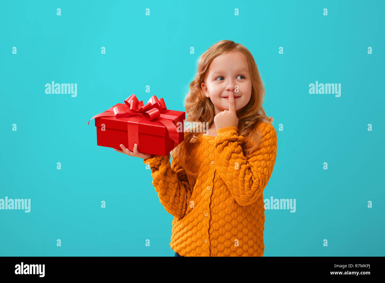 Portrait von ein kleines Mädchen in einem gestrickten Pullover auf einem cyan Hintergrund. Das Kind Träume nachdenklich seinen Finger auf seine Lippen und hält ein rotes Feld Stockfoto