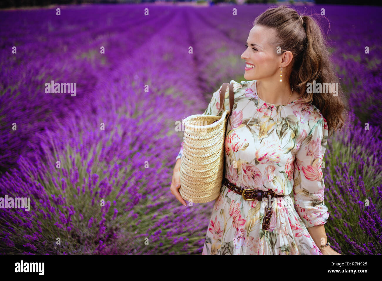 Happy Elegant Frau mit Stroh Beutel schauen in die Ferne in lavendelfeld in der Provence, Frankreich. Frau genießen Sie die endlosen Felder der blühenden Lave Stockfoto