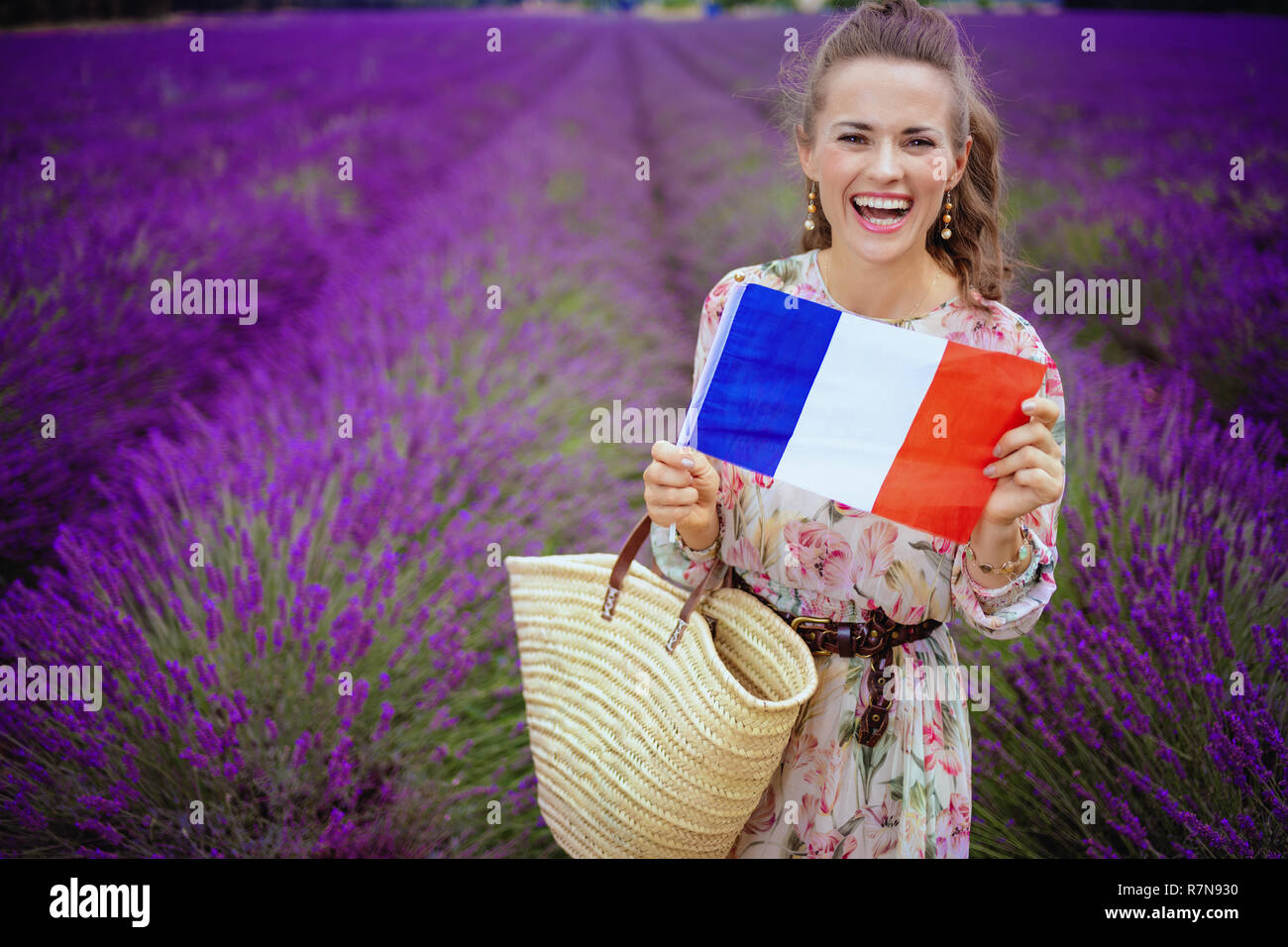 Lächelnd modernen Frau in langen Kleid mit Stroh Beutel, die französische Flagge im lavendelfeld in der Provence, Frankreich. Die romantische Anziehungskraft von Frankreich - Stockfoto