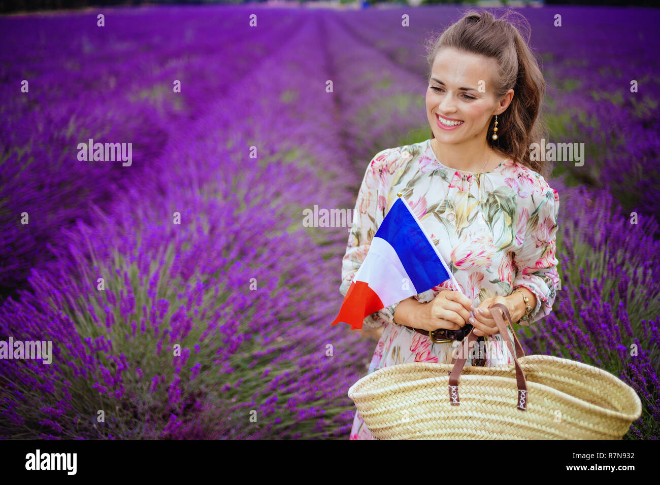 Happy Elegant Frau in geblümten Kleid mit Stroh Beutel und französische Flagge gegen Lavendelfeld in der Provence, Frankreich. Der Traum wurde wahr! Touristische Frau enjo Stockfoto