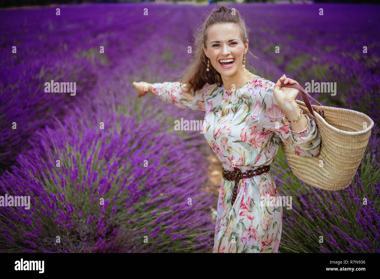 Lächelnd elegante Frau in langen Kleid mit Stroh Beutel zeigen auf etwas gegen Lavendelfeld in der Provence, Frankreich. Provence gefällt Romantiker mit s Stockfoto