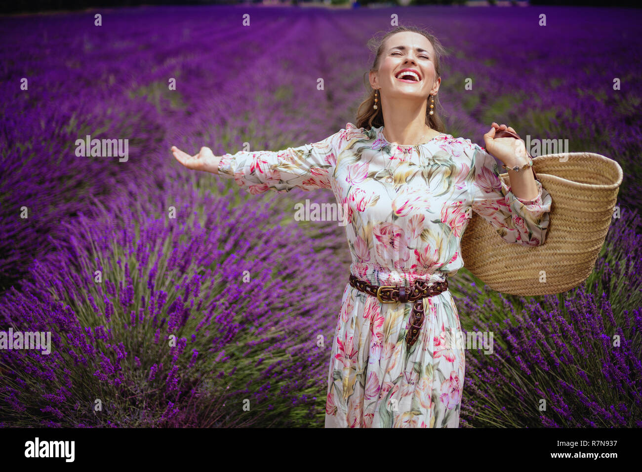 Happy Elegant Frau in geblümten Kleid mit Stroh Beutel voller Freude an lavendelfeld in der Provence, Frankreich. Erstaunlich Lila Lavendelfelder der Provence sind ein s Stockfoto