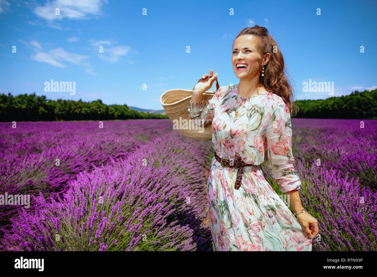 Lächelnde junge Frau mit Stroh Beutel Suche in Entfernung gegen Lavendelfeld in der Provence, Frankreich. Die Natur der Provence und der magische Duft von laven Stockfoto