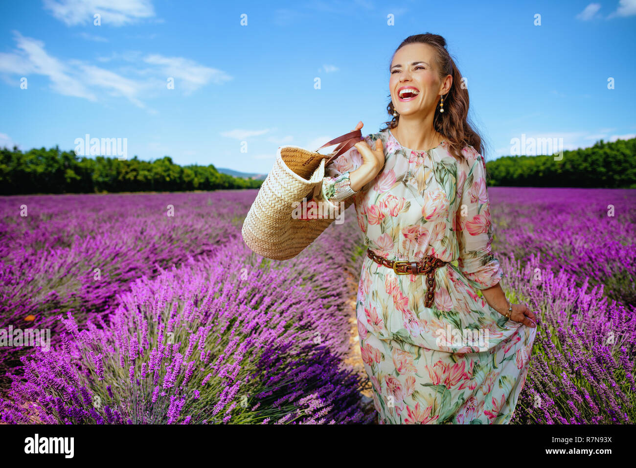 Gerne trendy Frau in geblümten Kleid mit Stroh Beutel schauen in die Ferne gegen Lavendelfeld in der Provence, Frankreich. Sommer in der Provence nicht le Stockfoto