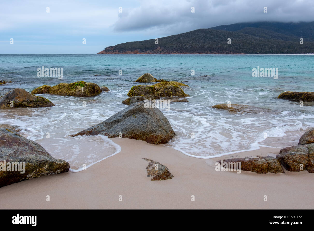 Strand am Wineglass Bay, Freycinet Nationalpark, Tasmanien mit Steinen in den Vordergrund und blaues Meer an einem bewölkten Tag Stockfoto