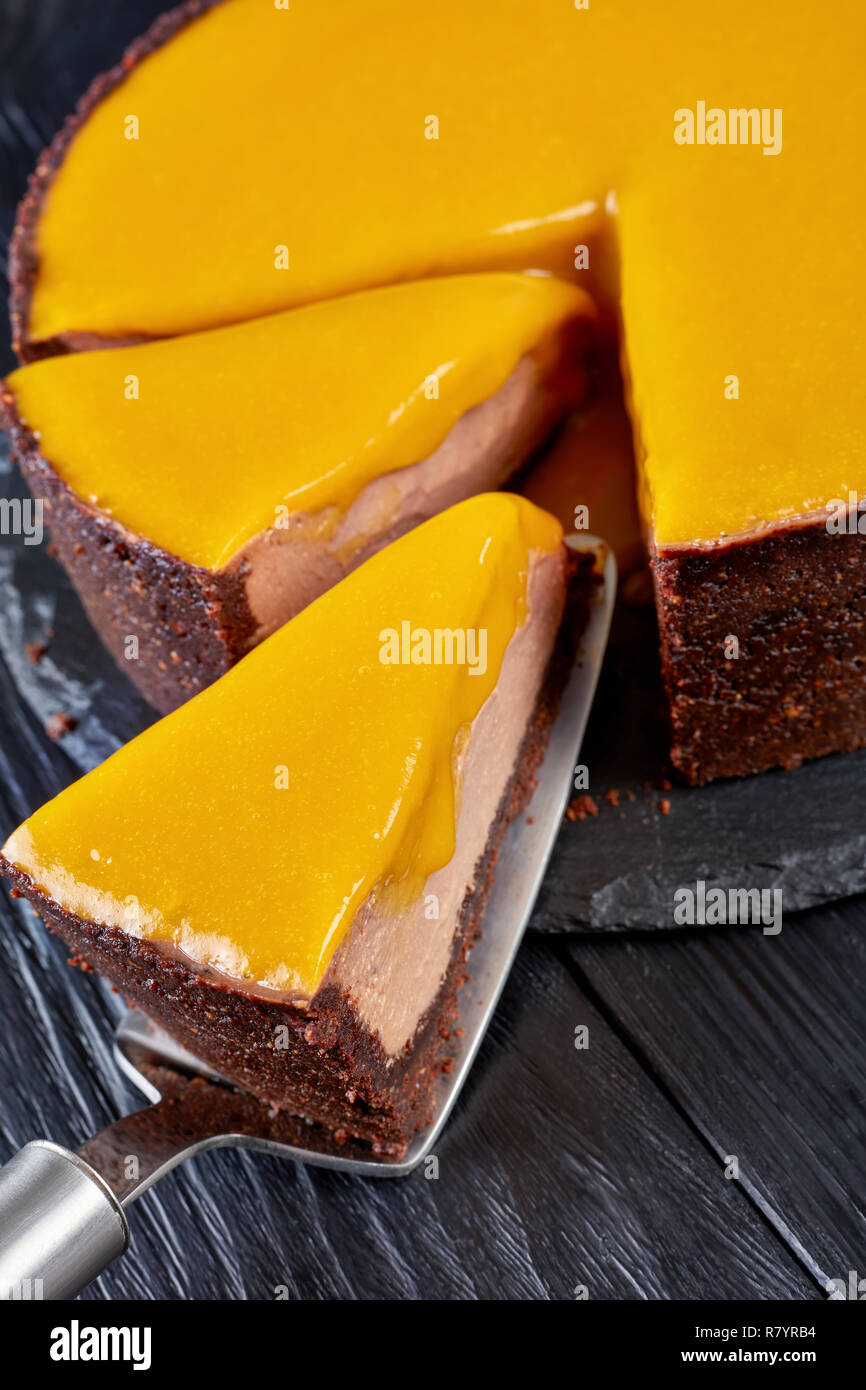 In Scheiben geschnittene Schokolade Mango cheesecake. ein Stück auf einem Kuchen Schaufel auf einen schwarzen Stein Platte auf einem schwarzen Holztisch, vertikale Ansicht von oben, close-up Stockfoto