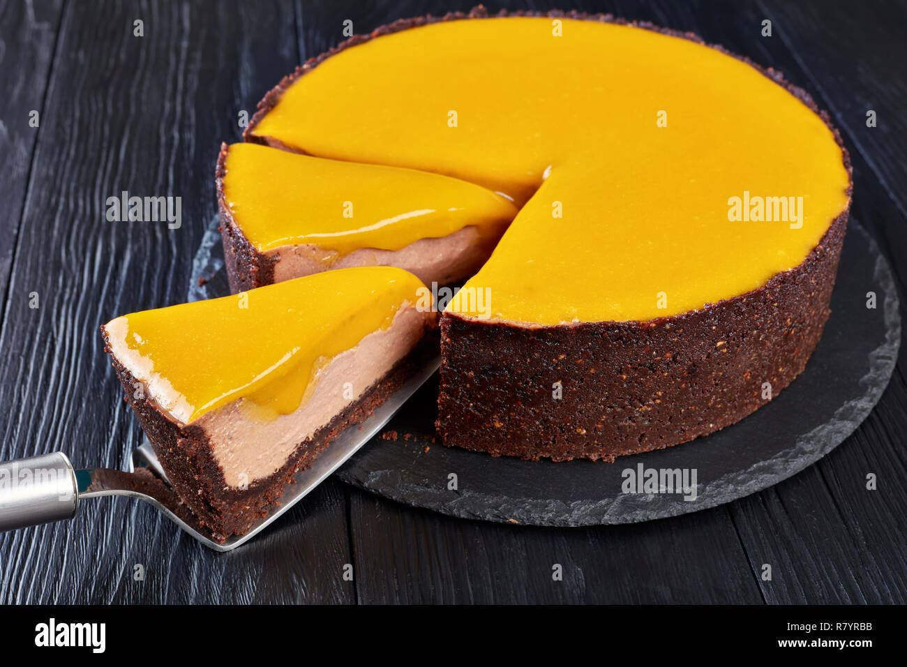 In Scheiben geschnittene Schokolade Mango cheesecake. ein Stück auf einem Kuchen Schaufel auf einen schwarzen Stein Platte auf einem schwarzen Holz- Tabelle, Ansicht von oben, close-up Stockfoto