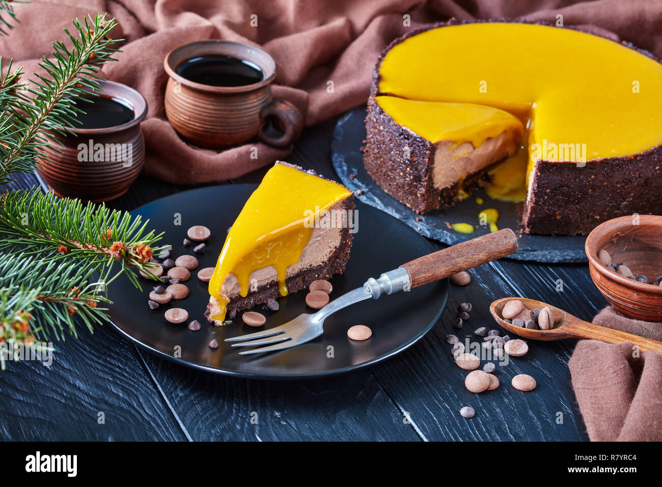 Close-up ein Stück Schokolade Mango cheesecake serviert auf einem schwarze Platte mit Dessert Gabel und Chocolate Chips. Tanne und Tassen mit Kaffee auf einem Stockfoto