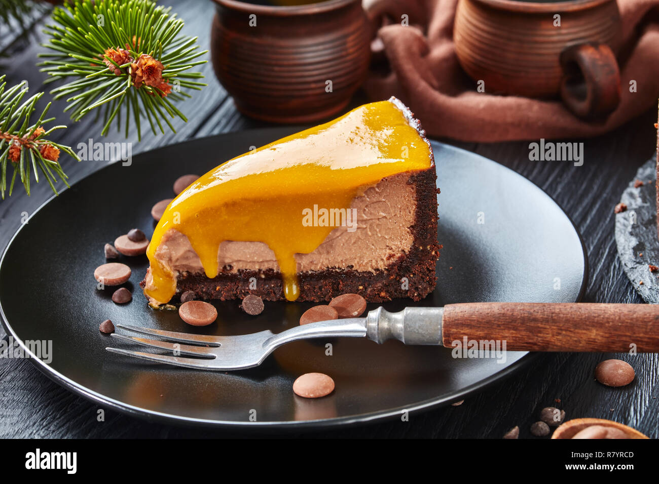 Close-up ein Stück Schokolade Mango cheesecake serviert auf einem schwarze Platte mit Dessert Gabel und Chocolate Chips. Tanne und Tassen mit Kaffee an. Stockfoto