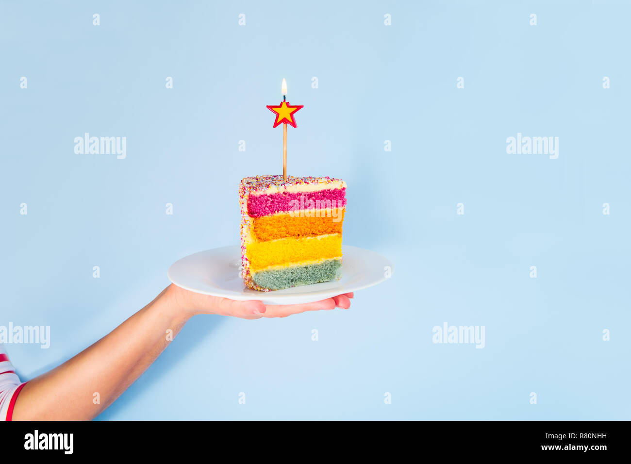 Weibliche Hand, die eine weiße Platte mit Slice von Rainbow Kuchen mit birning Kerze in der Form von Sternen auf blauem Hintergrund isoliert. Happy bithday, party Stockfoto