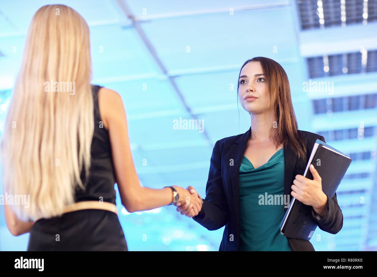 Zwei Mädchen im Büro mit Ordnern mit Papieren in der Hand. Junge Frauen Hände schütteln. Blaue leere Halle auf dem Hintergrund Stockfoto