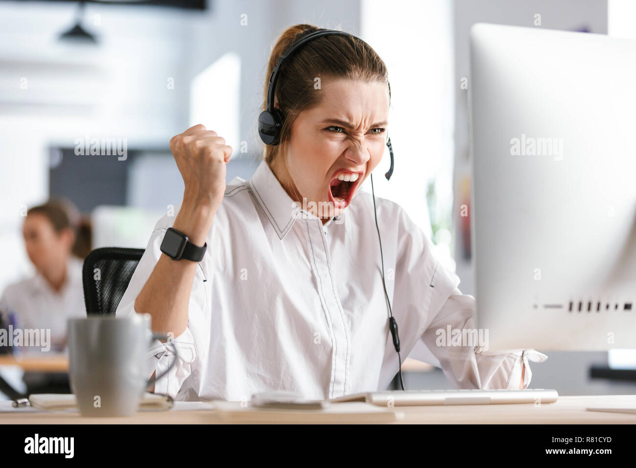 Wütende junge Frau mit Mikrofon Headset im Hemd an Ihrem Arbeitsplatz im Büro sitzen, gekleidet, schreien Stockfoto