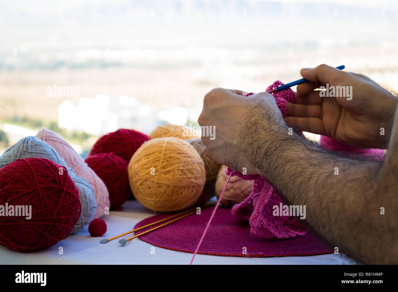 Ein Mann strickt ein rosa Schal für seine Tochter im Freien auf einem hellen, sonnigen Tag. Nahaufnahme der behaarten Händen eines Mannes. Tag der Frau. Stockfoto