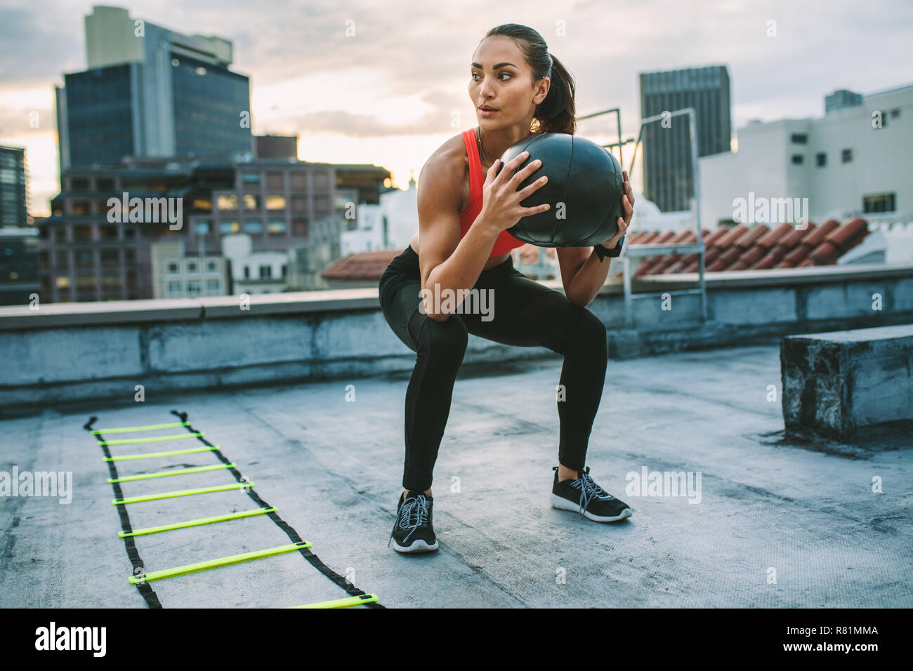 Fitnesstraining-Frau auf dem Dach mit einem Medizinball. Frau, die Squats macht, hält einen Medizinball mit einer Agilitätsleiter an ihrer Seite auf dem Dach Stockfoto