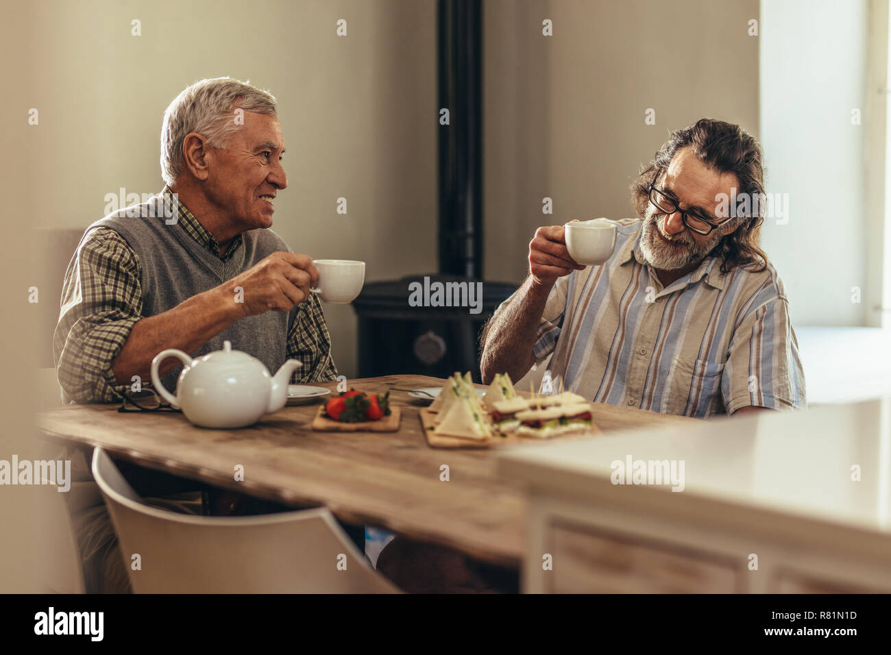 Zwei ältere Männer, die Kaffee Tassen beim zusammen am Tisch drinnen sitzen. Pensionierte Männer genießen Sie Kaffee und Snacks zusammen zu Hause. Stockfoto