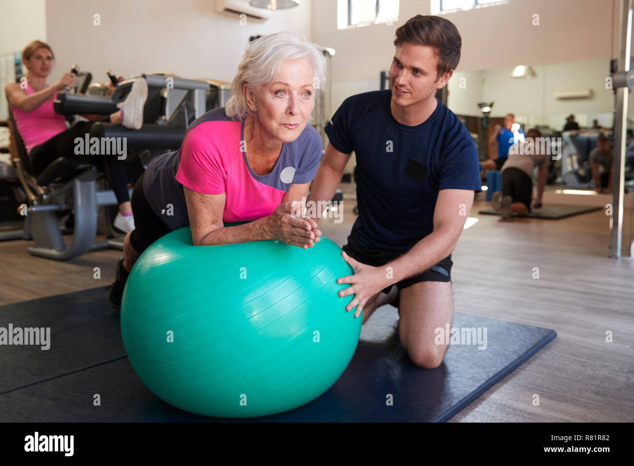 Ältere Frau Ausübung auf Schweizer Kugel durch Personal Trainer im Fitnessstudio gefördert Stockfoto