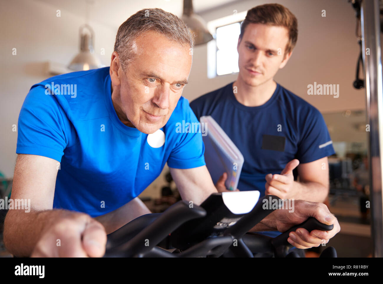 Älterer Mann Ausübung auf Radfahren Maschine vom persönlichen Trainer im Fitnessstudio gefördert Stockfoto