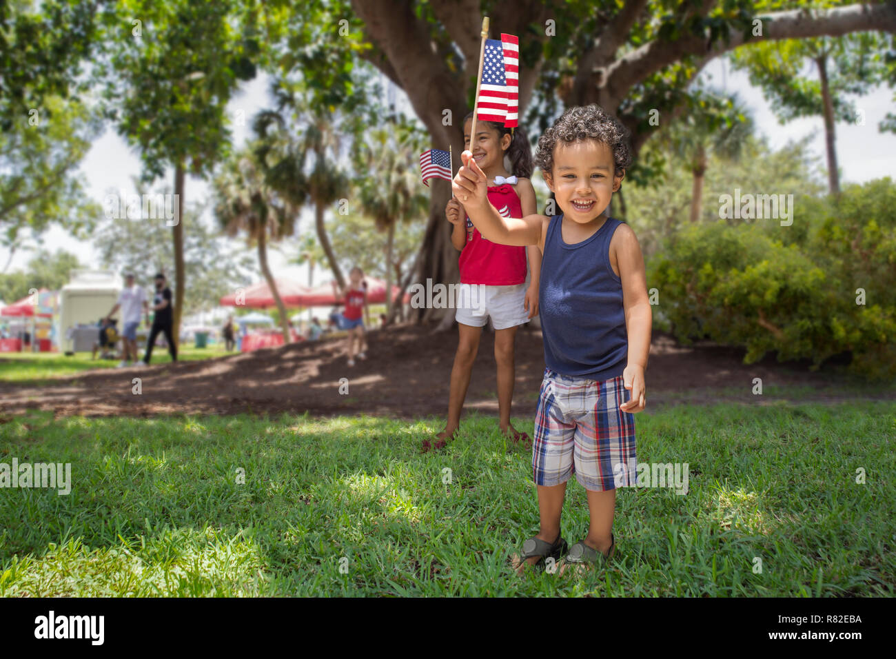 Hispanic Bruder und Schwester spielen an der Community Park im Schatten eines großen Baumes, während Sie die amerikanische Flagge stolz halten. Stockfoto