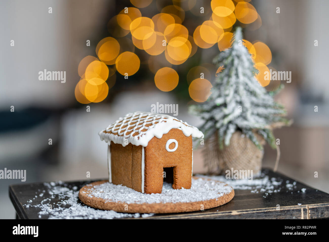 Lebkuchenhaus auf hölzernen Tisch. Defokussierten Girlande Lichter auf Hintergrund. Weihnachtsbaum und Urlaubsstimmung. Morgen im hellen Wohnzimmer. Stockfoto