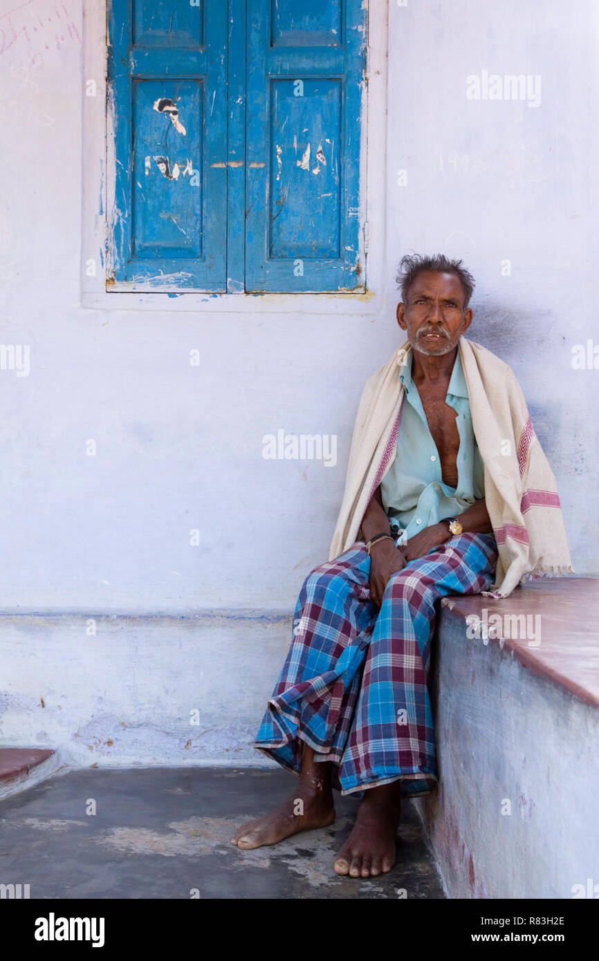 Ein alter Mann sitzt vor einem Haus in Tamil Nadu, Indien. Das Haus hat gemalte Putzwände in hellen Pastelltönen. Die Fensterläden sind blau. Stockfoto