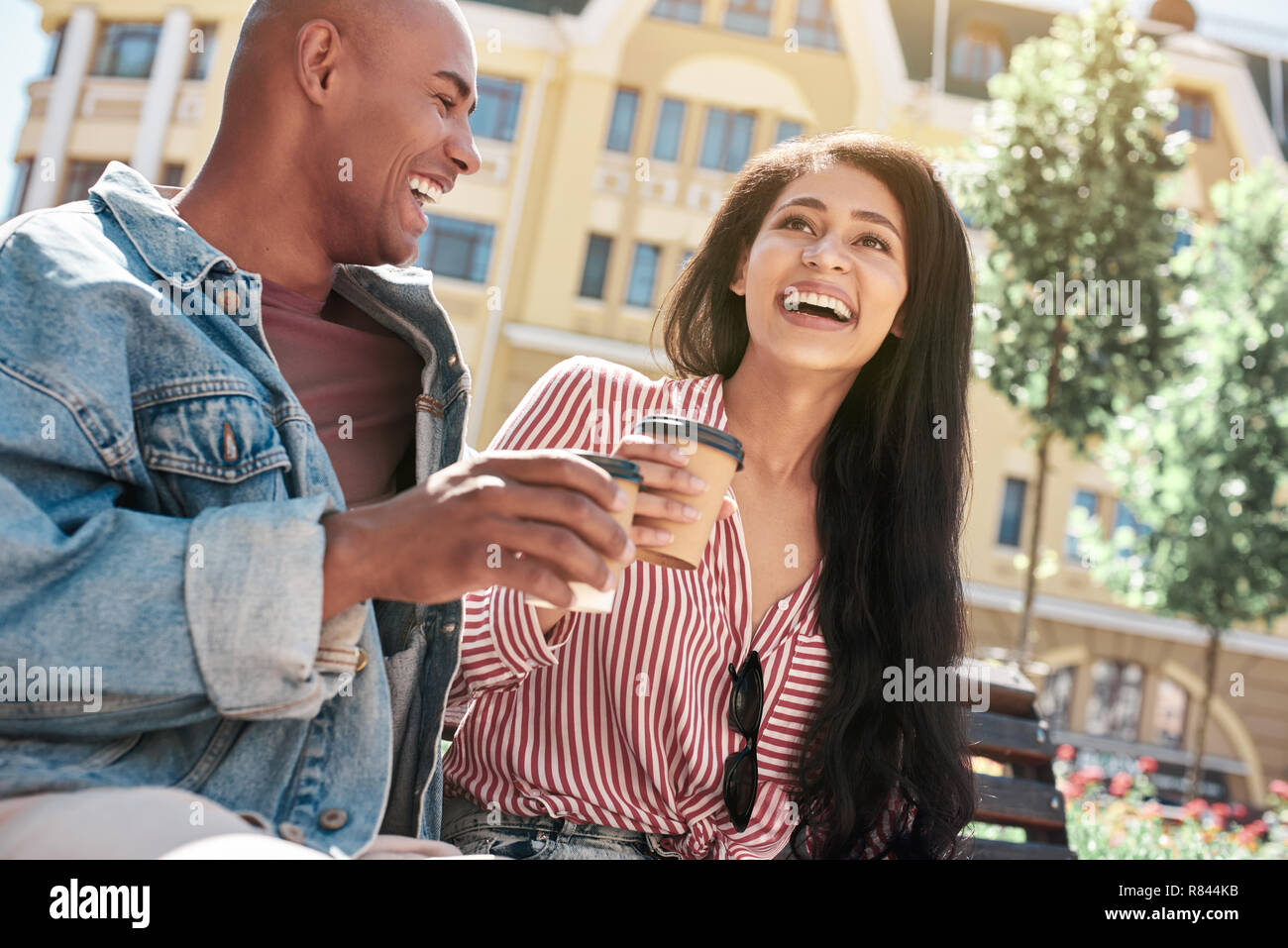 Romantische Beziehung. Junge diverse Paar sitzt auf der Bank auf die Stadt Straße holding Tassen trinken heißen Kaffee reden lachen Heiter close-up Stockfoto