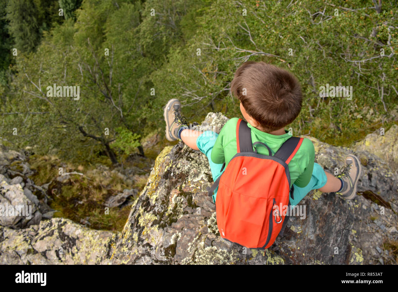 Junge sitzt auf einem felsigen Rand über einen Abgrund in die Berge, mit Bäumen sichtbar unterhalb, unscharf Stockfoto