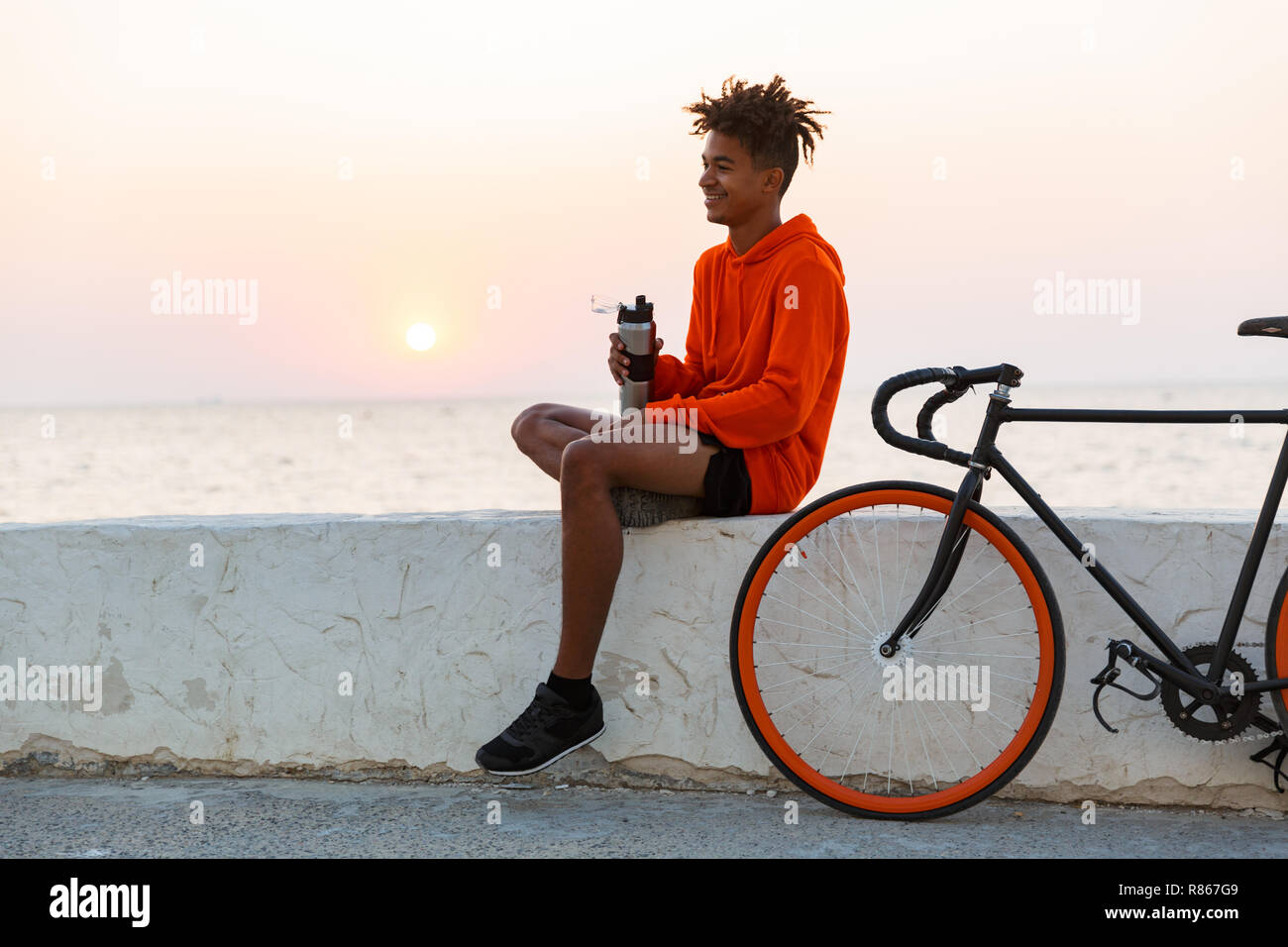 Bild von einem gutaussehenden jungen afrikanischen Mann mit Fahrrad im Freien am Strand Trinkwasser. Stockfoto