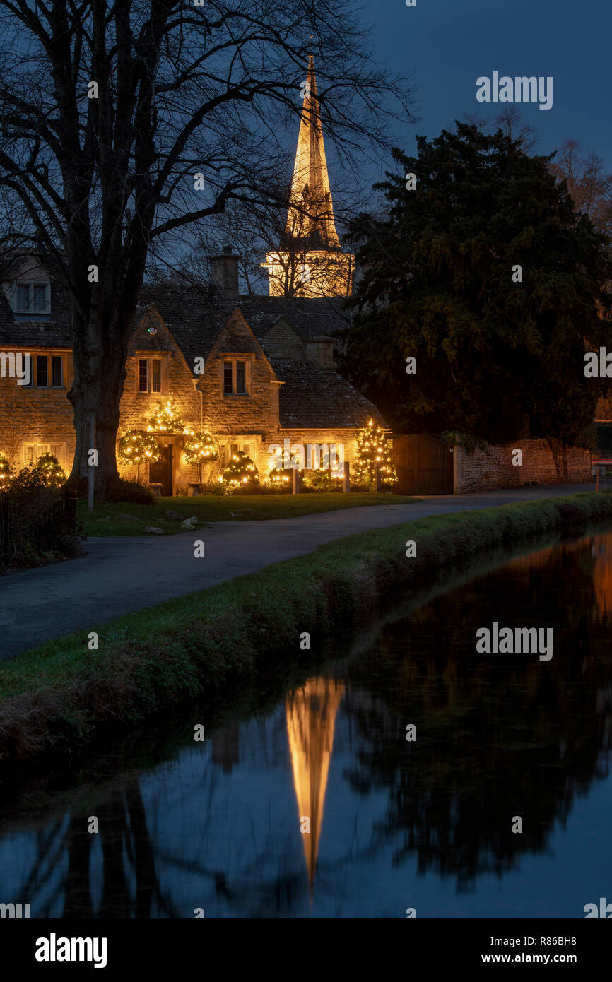 Weihnachtslichter auf einem cotswold-Häuschen mit der Kirche spiegeln sich in Lower Slaughter in der Nacht im Flussauge wider. Cotswolds, Gloucestershire, England Stockfoto