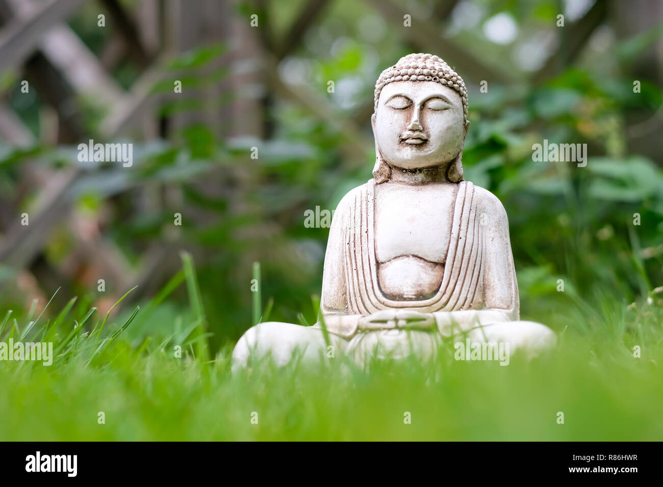 Kleine weiße Buddha Statue in einer Meditation pose mit grünem Gras im Vordergrund und die natürlichen hellen verschwommenen Hintergrund. Religiöses Symbol des Buddhismus Stockfoto