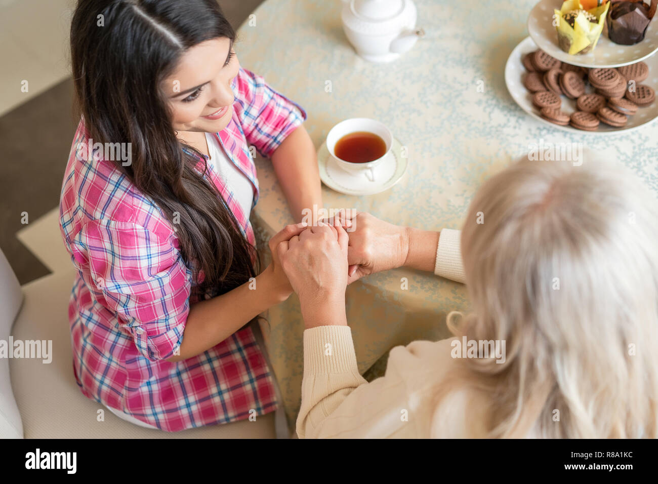 Aufnahme von oben auf eine hübsche junge Tochter Sitzen am Tisch bei einer Tasse Tee und Keksen und Hände mit Ihrer betagten Mutter. Stockfoto