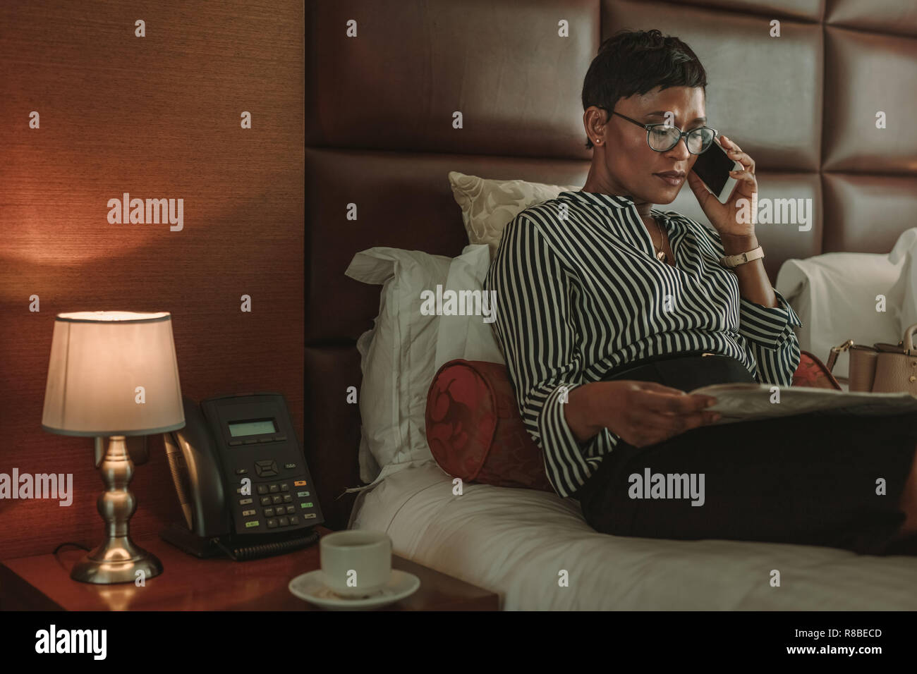 Reifen afrikanischen Geschäftsfrau auf Hotel Zimmer Bett und liest Zeitung und Gespräch am Handy. Afrikanische Frauen in formelle Kleidung Entspannung auf Bett mit Nachrichten Stockfoto