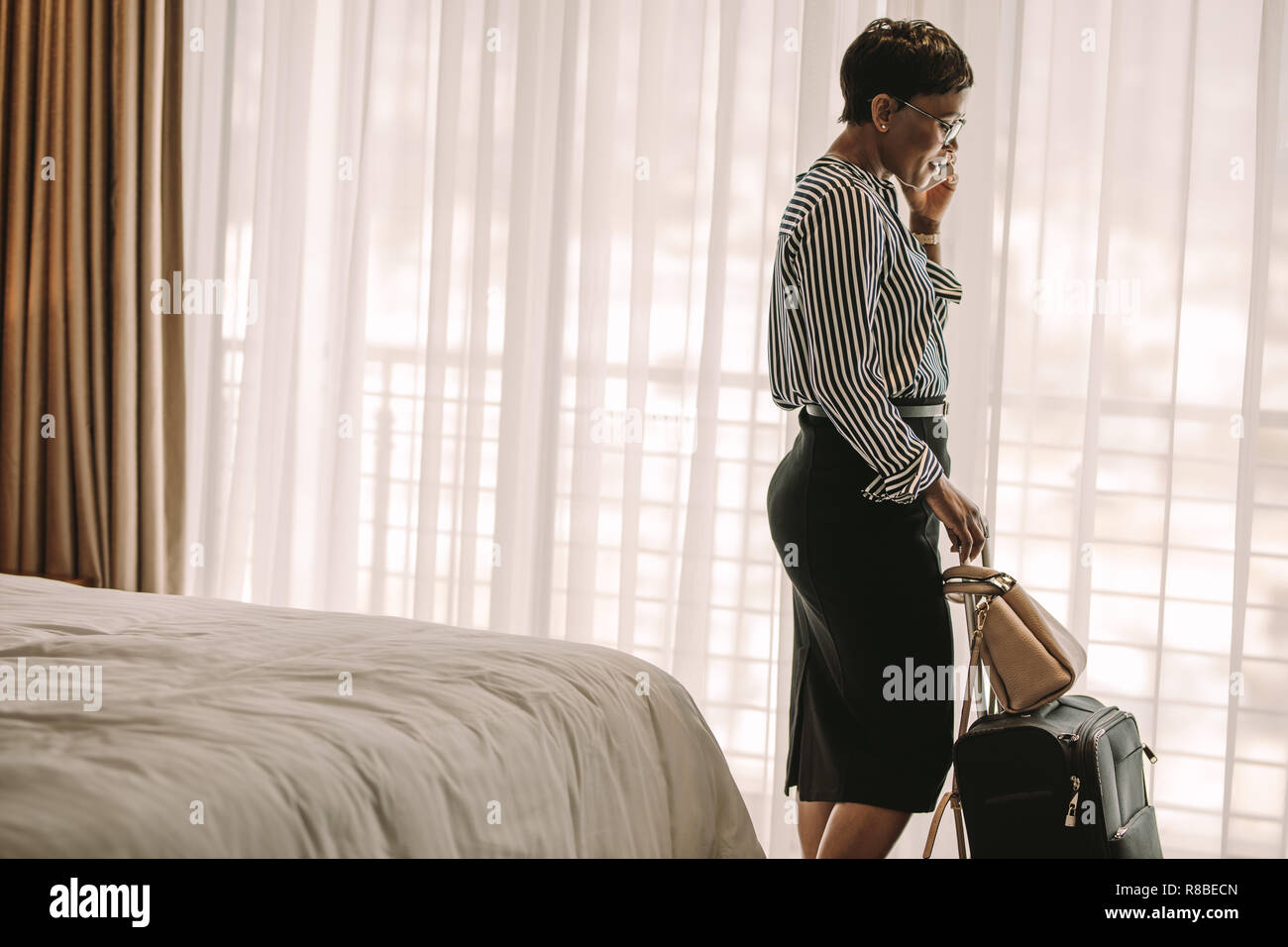 Frau in formelle Kleidung durch Bett im Hotelzimmer mit ihrem Gepäck Sprechen über Handy stehen. Weibliche CEO auf Geschäft Tour am Telefon sprechen von Hote Stockfoto