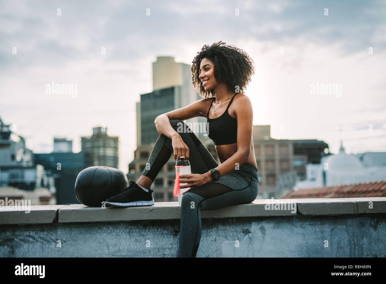 Seitenansicht einer Sportlerin sitzen auf der Dachterrasse Zaun mit einem medizinball Trinkwasser nach dem Workout. Lächelnde Frau in Fitness Kleidung entspannende aft Stockfoto