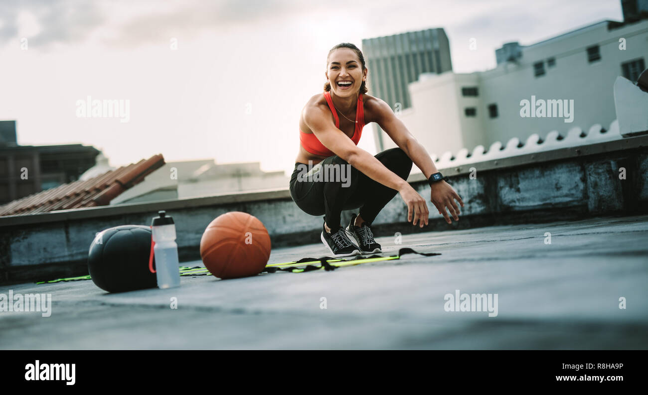 Fitness-Frau sitzt auf den Zehen, während sie auf dem Dach trainiert. Fröhliche Athletin, die auf dem Dach mit Medizinball und Basketball trainiert Stockfoto