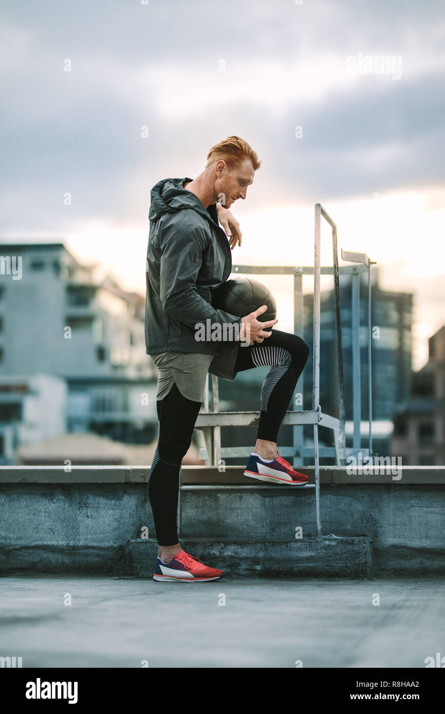 Fitness-Mann, der auf dem Dach steht und einen Medizinball hält. Seitenansicht eines entspannten Athleten, der in der Nähe einer Dachtreppe steht und nach unten blickt. Stockfoto