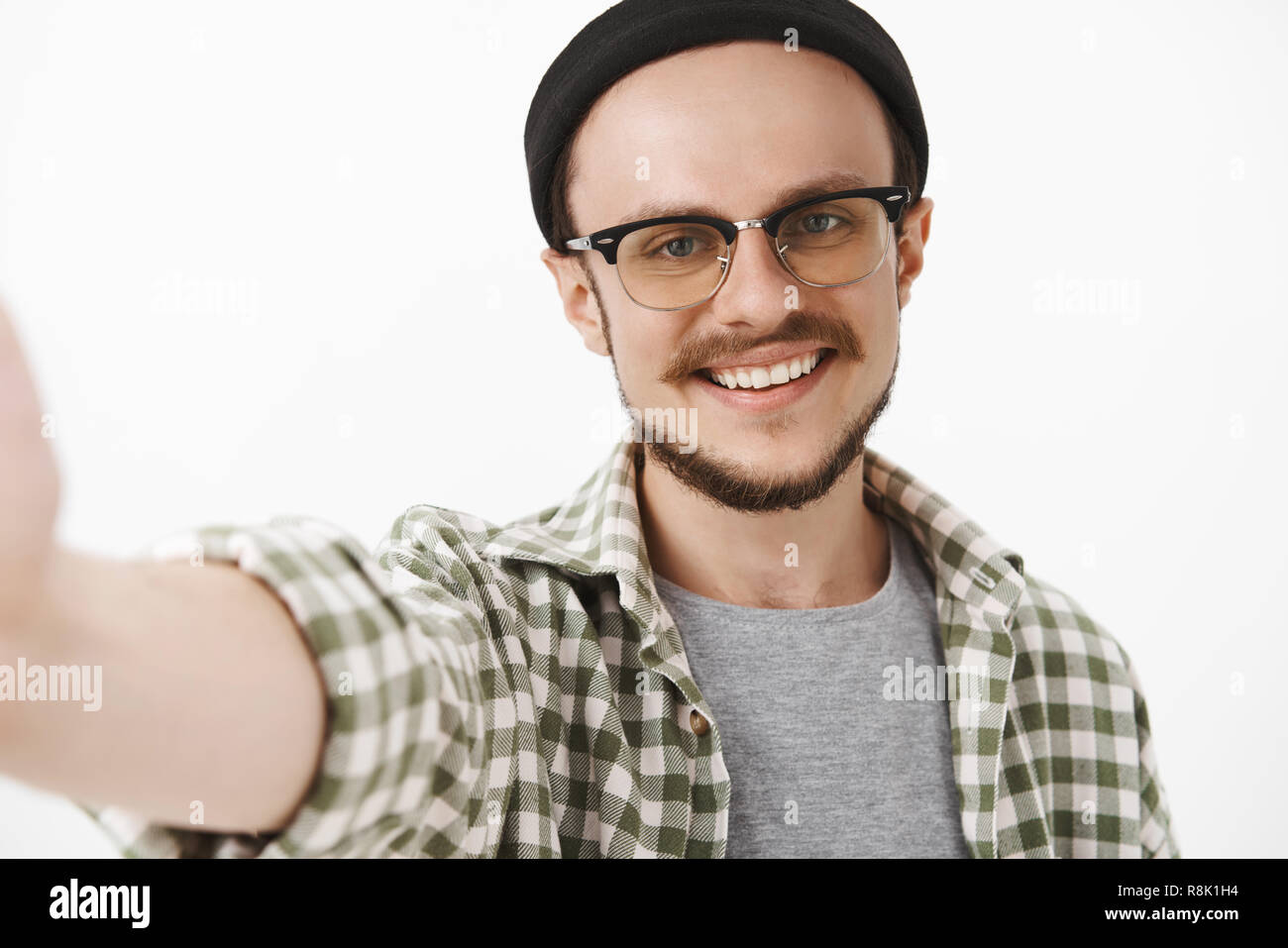 Nahaufnahme der freundlich schön ordentlich Kerl in Schwarz trendige Mütze und Brille mit Schnurrbart breit grinsend Hand ziehen Kamera zu halten, während der Einnahme von selfie auf neues Smartphone Stockfoto