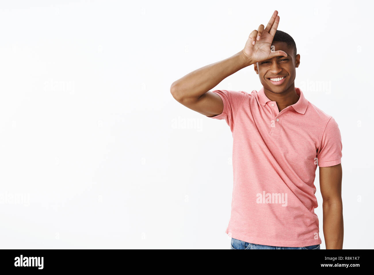 Verlierer zu Hause bleiben. Freundlich, fröhlich und selbstbewussten jungen afrikanischen amerikanischen männlichen Kursteilnehmer in Pink polo shirt, Buchstaben L auf der Stirn als spöttisch Freund letzten, posieren heiter über graue Wand Stockfoto