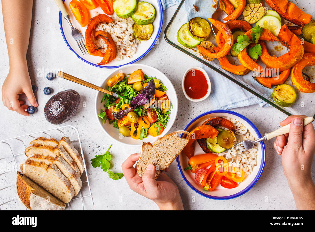 Flach der Familie Hände essen gesund essen. Vegan Mittagstisch Ansicht von oben. Gebackene Gemüse, frische Salate, Beeren, Brot auf einem weißen Hintergrund. Stockfoto