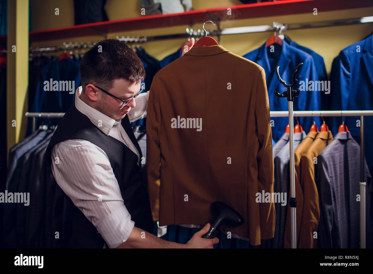 Individuelle Schnitte und näht einen Anzug. Ein junger Mann in der Schneiderei shop macht die Kleidung der Männer. Maßgeschneiderte Bekleidung Werkstatt. hält in ihrer Hand ein elektrisches Bügeleisen. wet - Wärmebehandlung Stockfoto