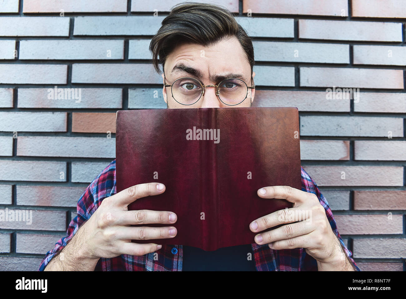 Lustiges Gesicht. Junge hipster Kerl tragen Brillen mit Abgeholt eine browe an Kamera suchen und halten ein Buch oder Buch auf Mauer Hintergrund. Stockfoto