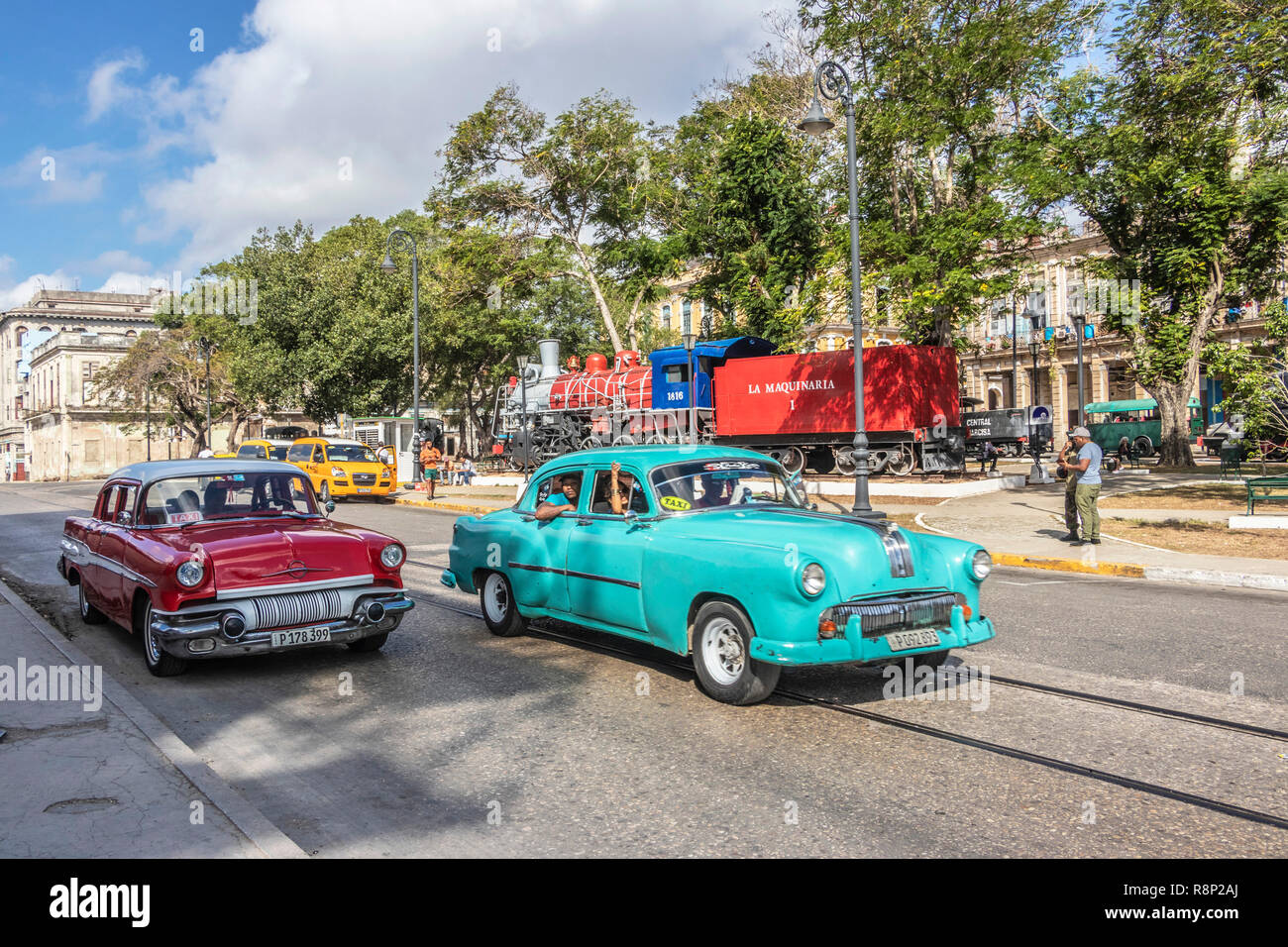 Oldtimer fahren entlang einer Straße in Havanna, Kuba, mit farbenfrohen Gebäuden und einer Zugausstellung im Hintergrund Stockfoto