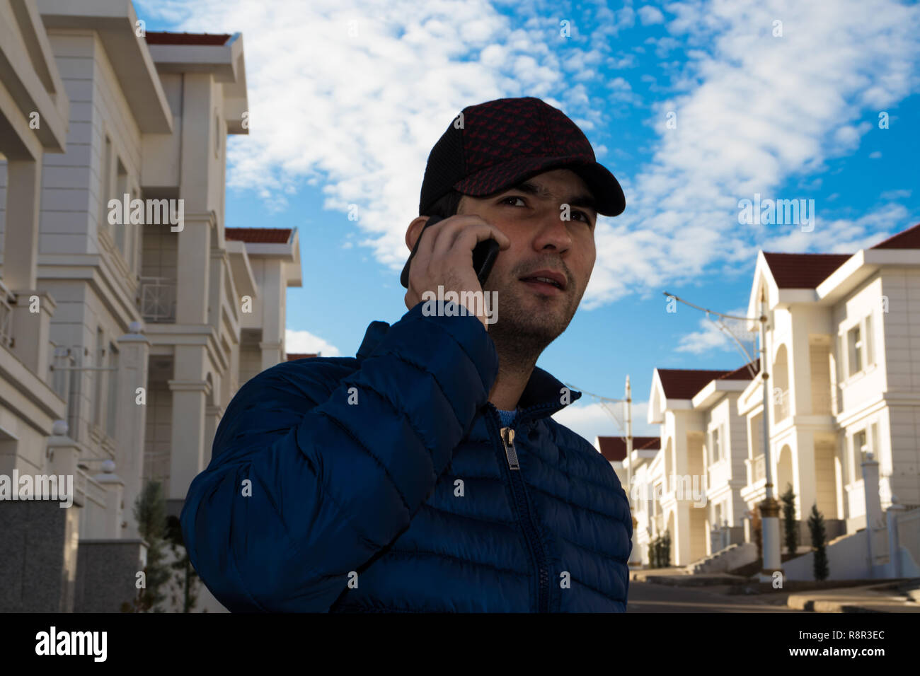 Immobilienmakler vor Villen in einem Gespräch per Handy mit einem potenziellen Kunden auf einem Hintergrund von einem blauen bewölkten Himmel Stockfoto
