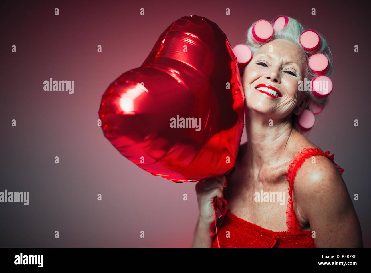 Portrait verspielte ältere Frau mit Lockenwicklern im Haar halten Herz-form Ballon Stockfoto