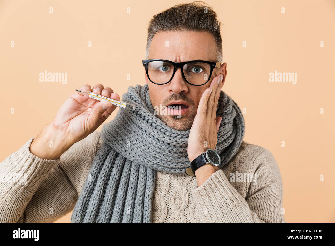 Porträt eines kranken Mann in Pulli und Schal stehend über beigen Hintergrund isoliert gekleidet, mit Thermometer Stockfoto