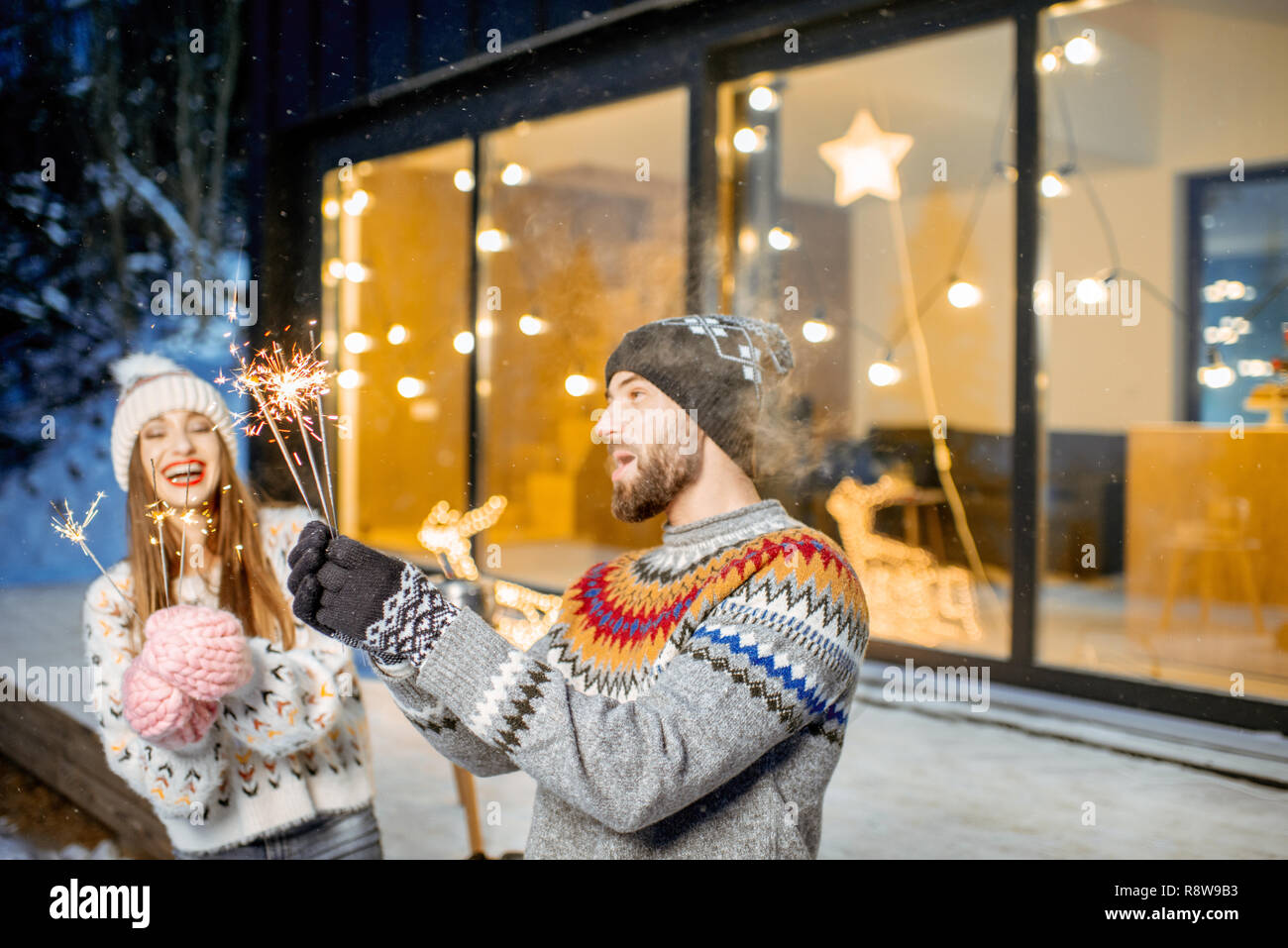 Junge glückliches Paar in Pullover feiern Winterurlaub mit bengalischen Feuer vor einem wunderschönen eingerichteten Haus gekleidet Stockfoto