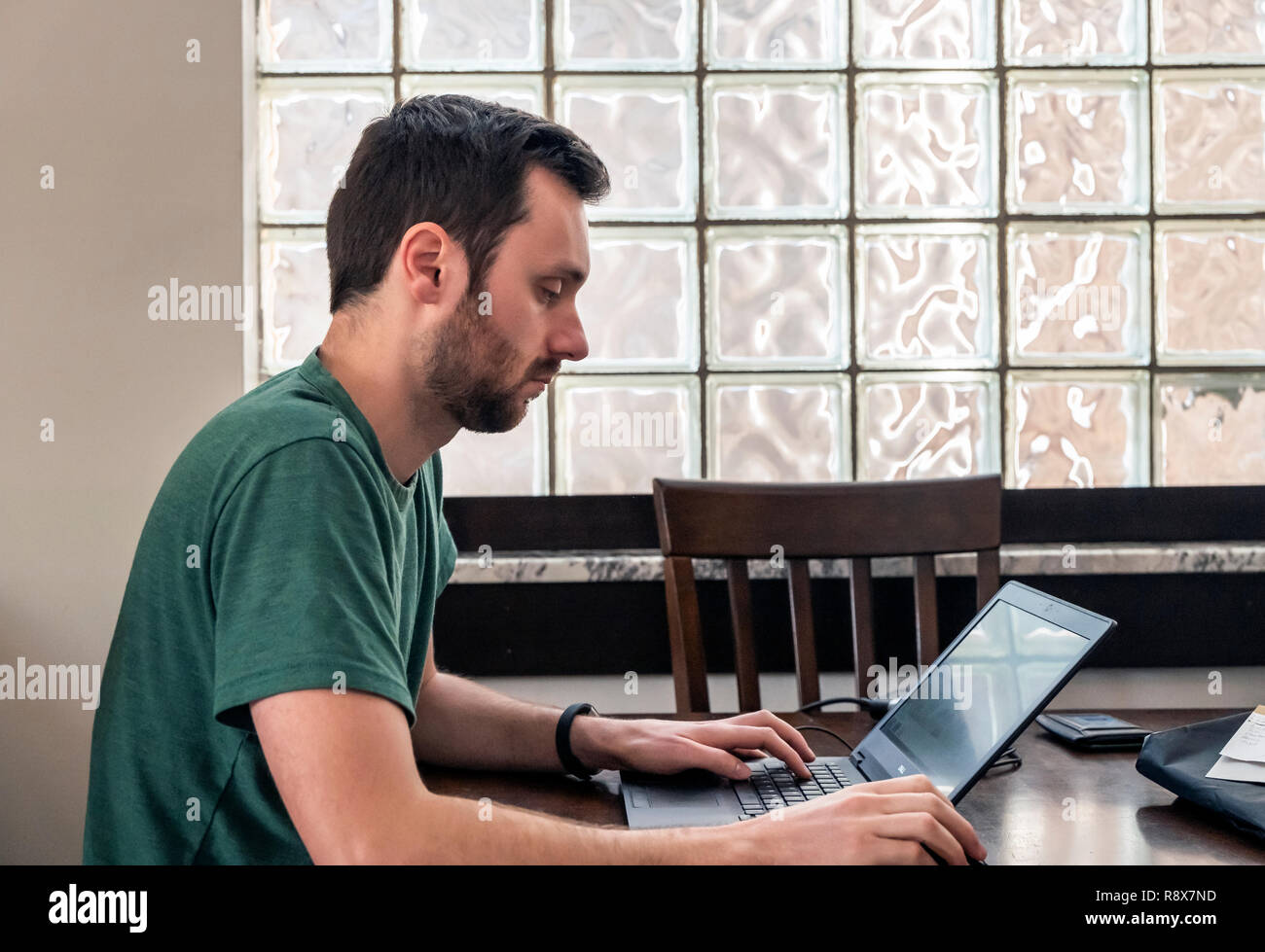 Junger bärtiger Mann zu Hause per Fernzugriff arbeiten am Laptop vor Glas Block Wand Stockfoto