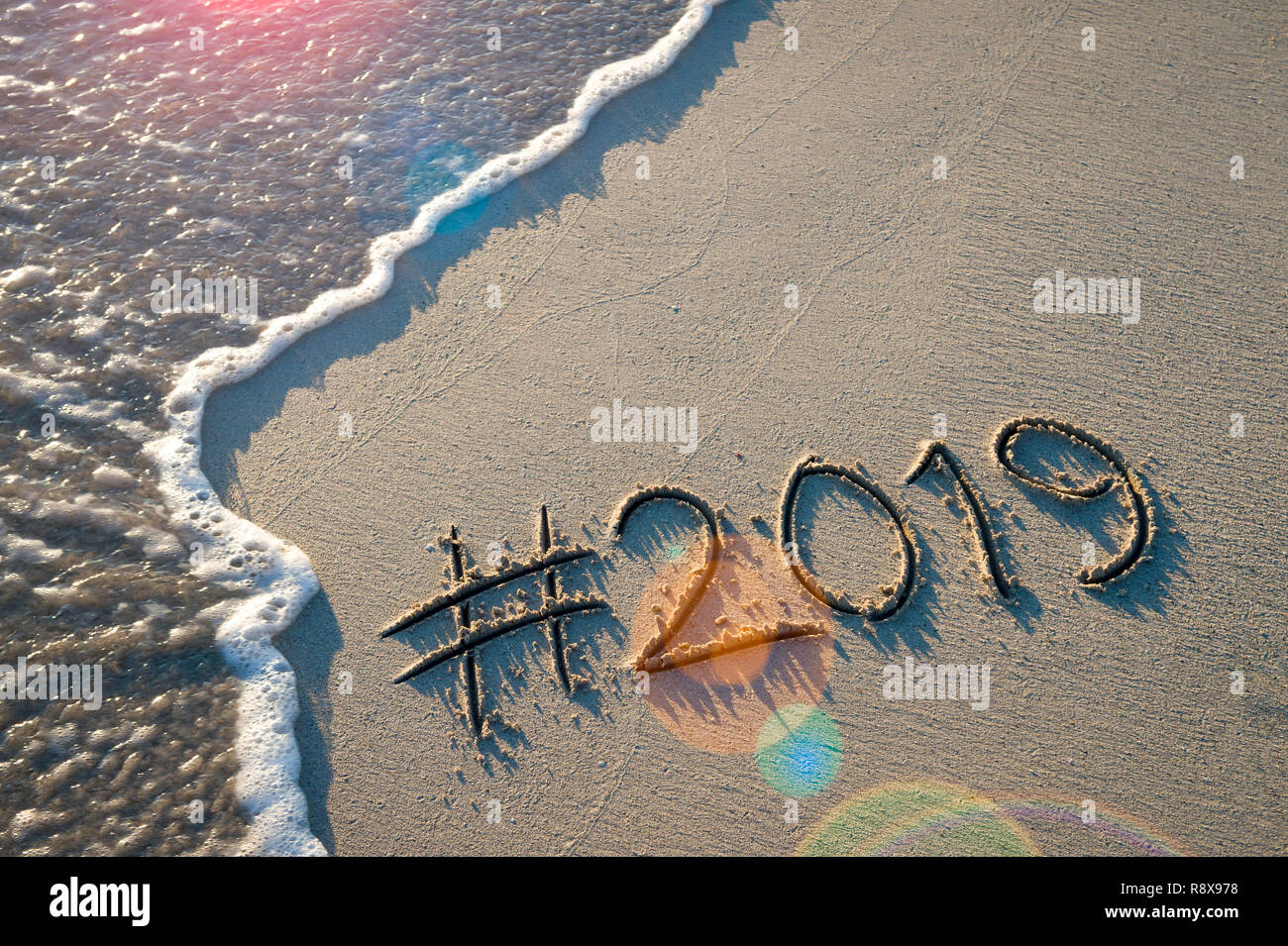 Modernes neues Jahr Nachricht mit social media Hashtag für 2019 handschriftlich in sanften Sand mit Sunrise lens flare über entgegenkommende Welle auf den Strand Stockfoto
