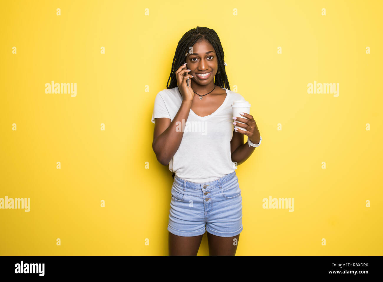 Attraktive dunkelhäutige Mädchen mit Afro Frisur tragen weiße T-Shirt in Telefongespräch mit ihrem Freund und trinken Kaffee isoliert auf Gelb Stockfoto