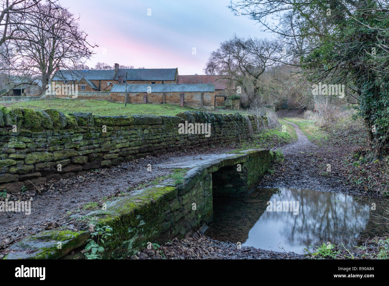 Unter einer Rosa vor Sonnenaufgang Himmel ein Fußweg und Trockenmauer führen über eine kleine steinerne Brücke über einen Bach, auf der rechten Seite ist ein Ford. Stockfoto