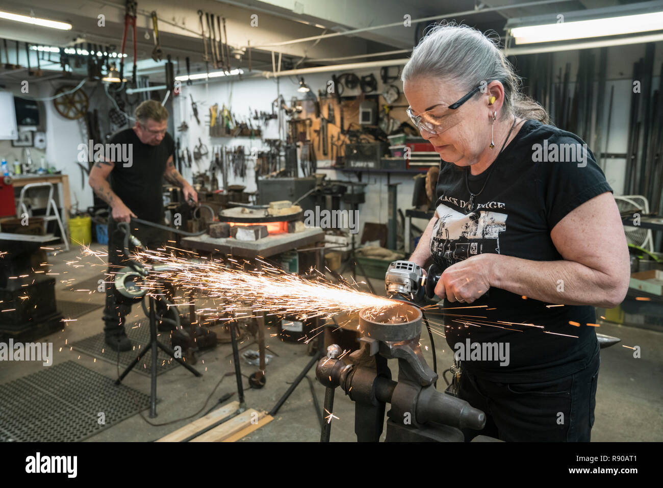 Kaukasische Frau schmied Schleifen auf einem Metallteil in Ihrem Studio. Männliche Schmied bei der Arbeit im Hintergrund. Stockfoto